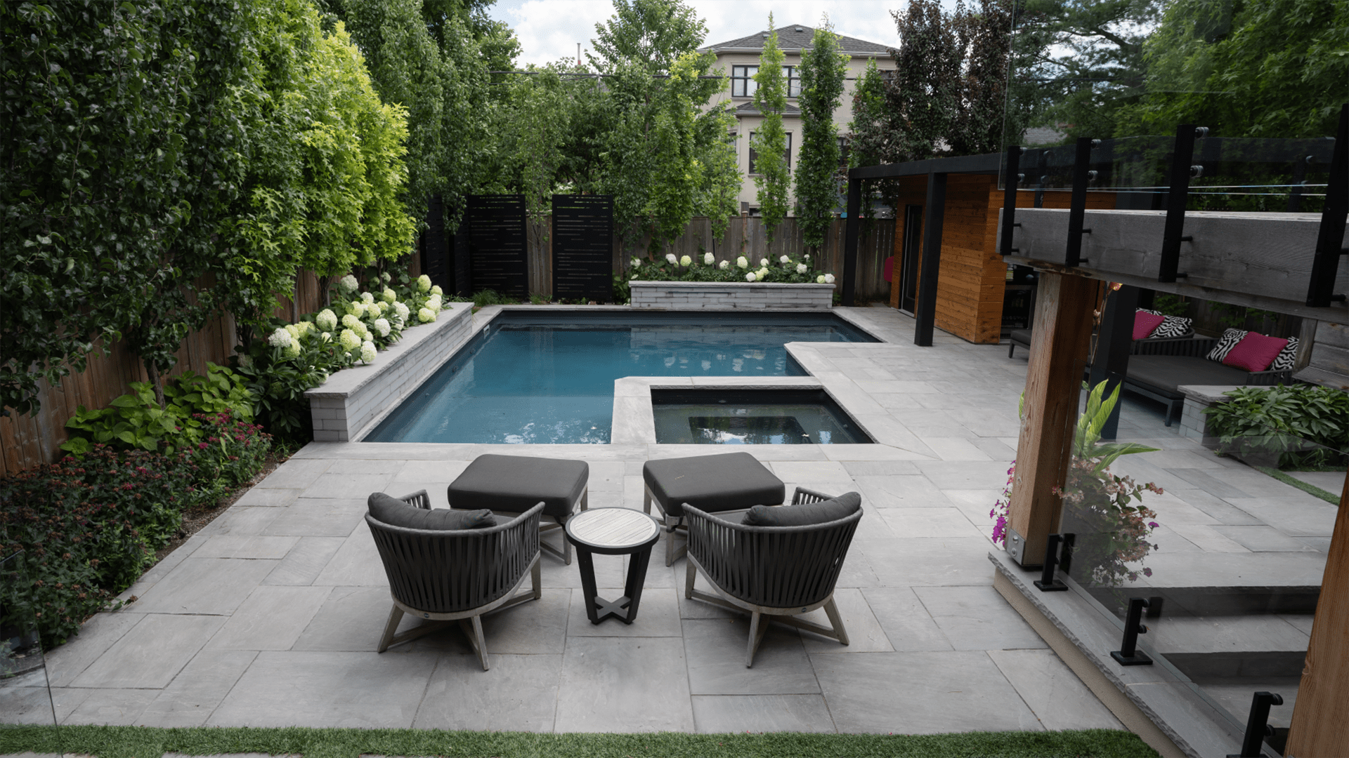 Modern backyard features a sleek pool, hot tub, and cozy seating area surrounded by lush greenery and privacy fences, ideal for relaxation.